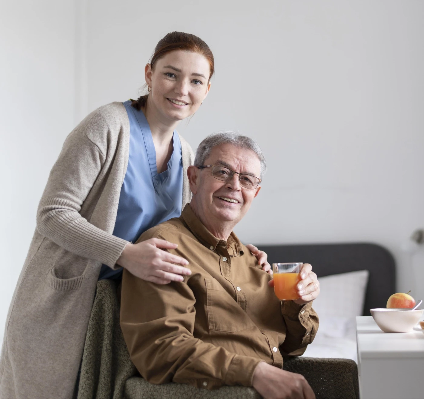 Pflegekraft lehnt sich lächelnd an einen Senior, der ein Glas Orangensaft hält